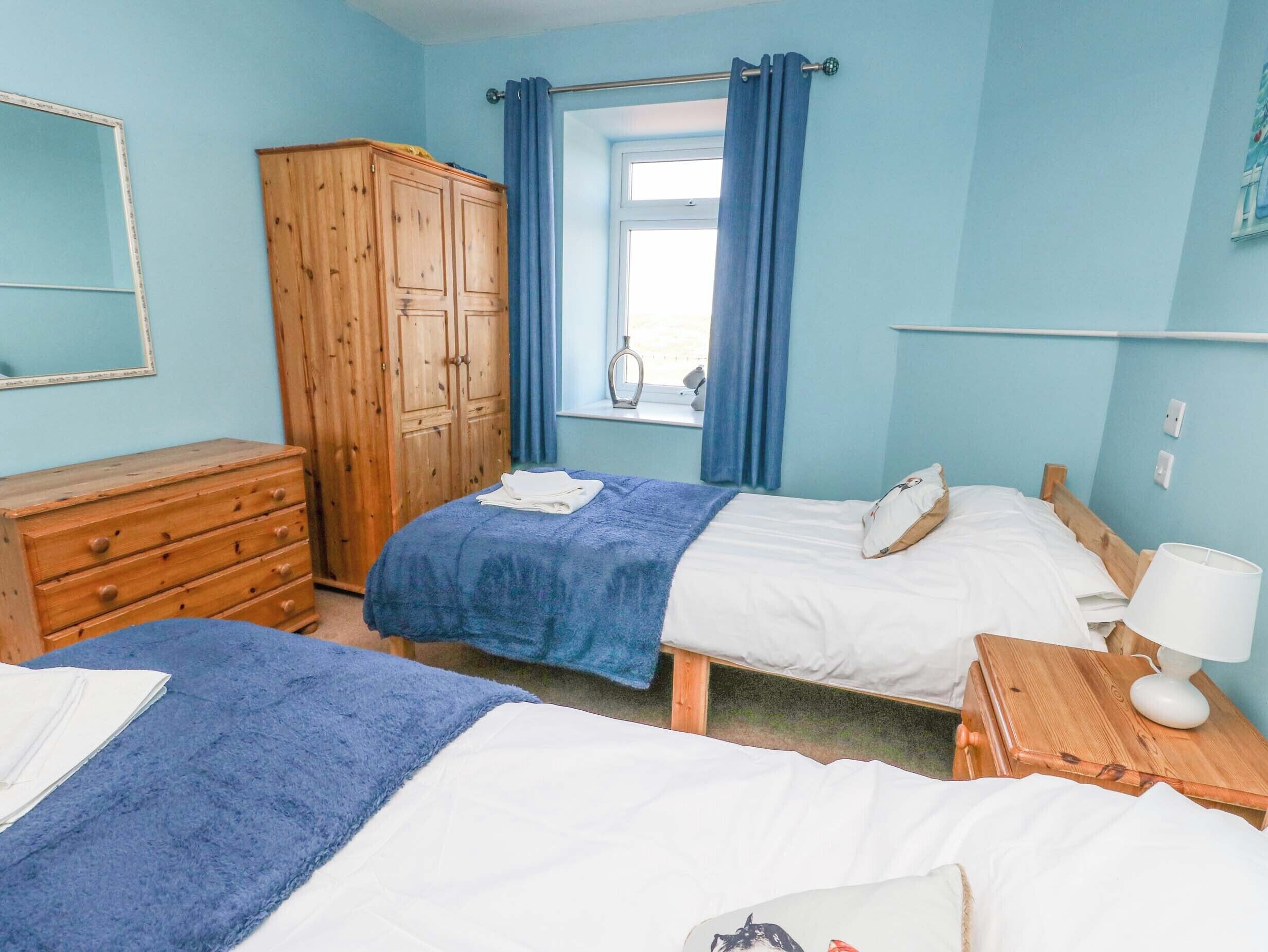 A bedroom with two beds, a wooden wardrobe, a window with curtains, and a bedside table with a lamp.