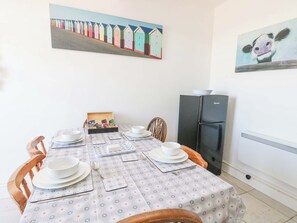 Un coin repas avec une table dressée pour quatre personnes, un réfrigérateur noir et un tableau représentant des cabanes de plage accroché au mur.