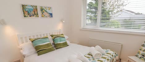 Una cama bien hecha con almohadas, cabecera blanca y vistas al exterior a través de una ventana con persianas.
