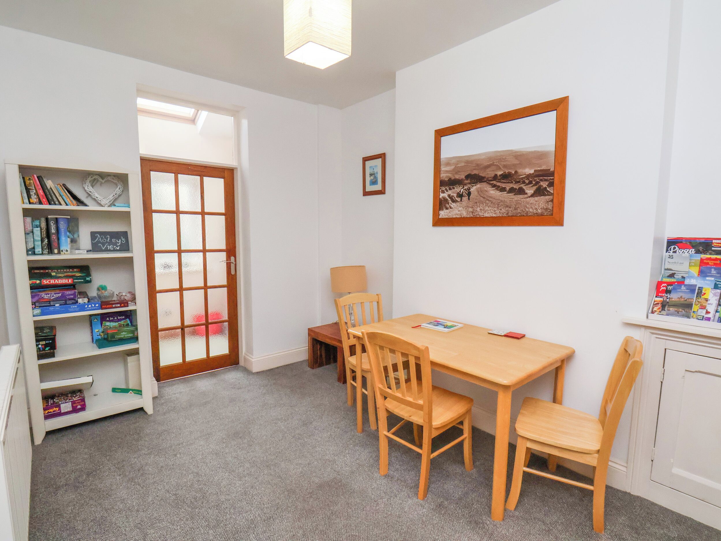 A room with a wooden table and chairs, a bookshelf with board games, a framed picture on the wall, and a door leading to another room.