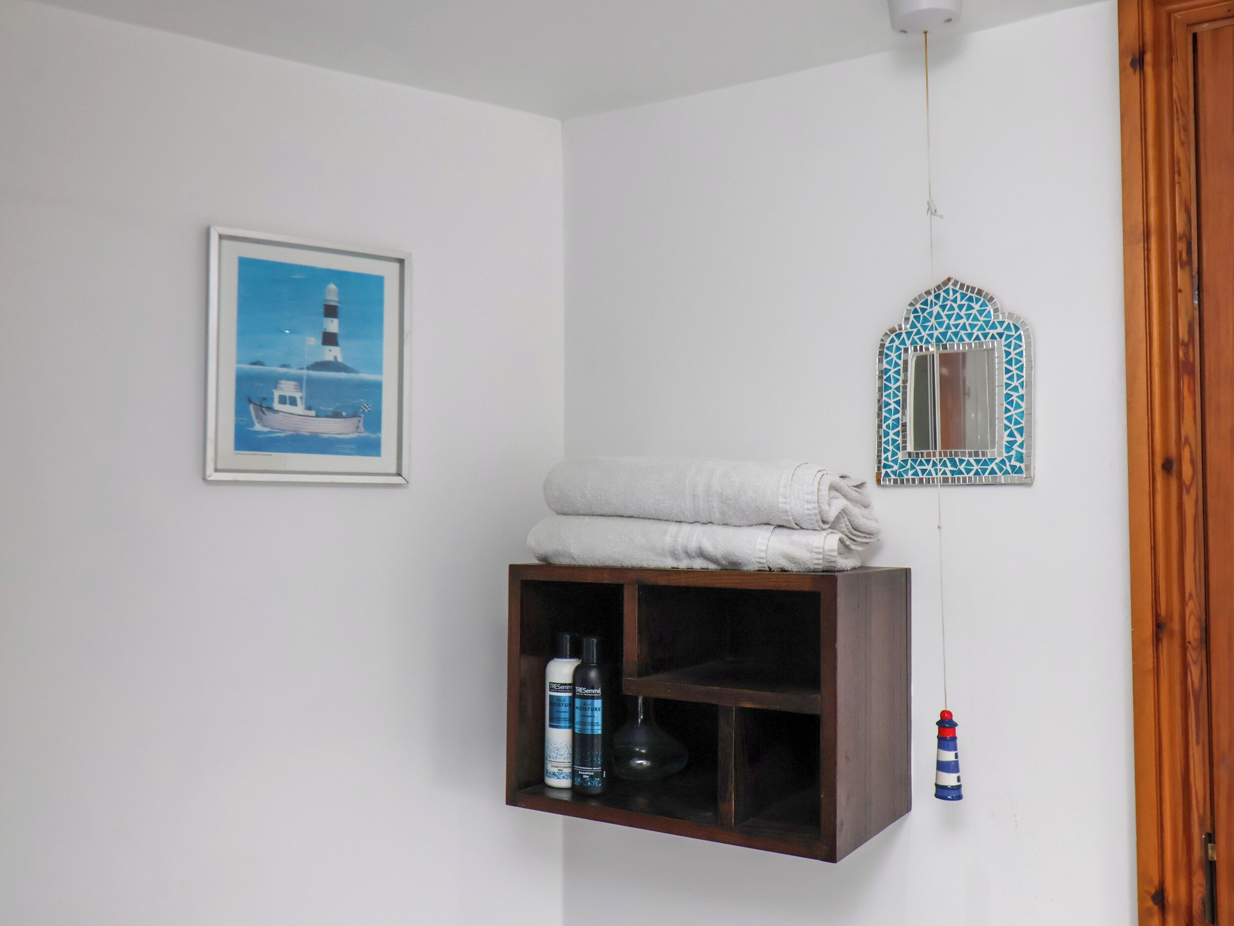 A bathroom with a framed picture of a lighthouse, a shelf with toiletries, and a decorative mirror.