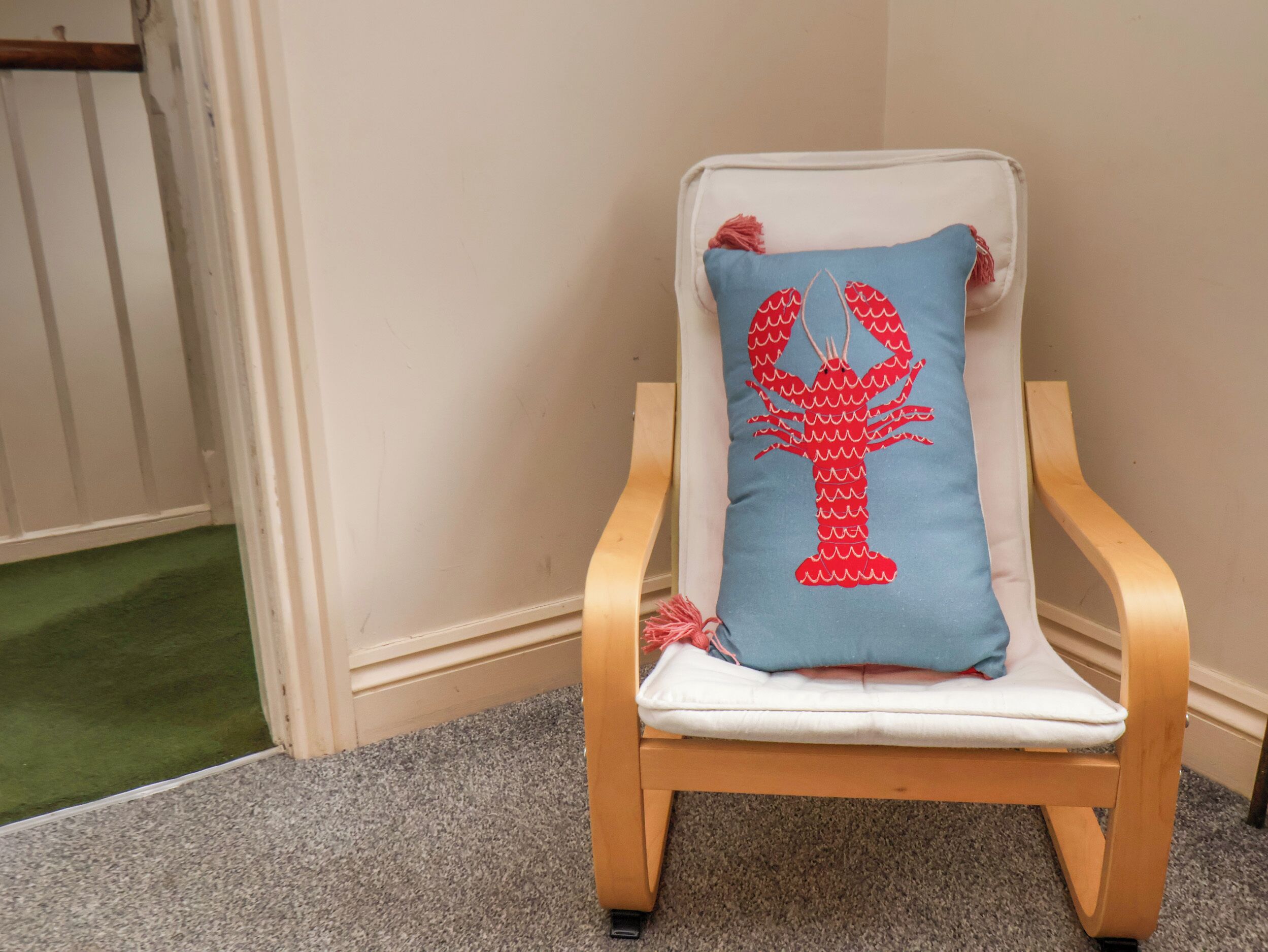 A wooden chair with a white cushion and a blue pillow featuring a red lobster design.