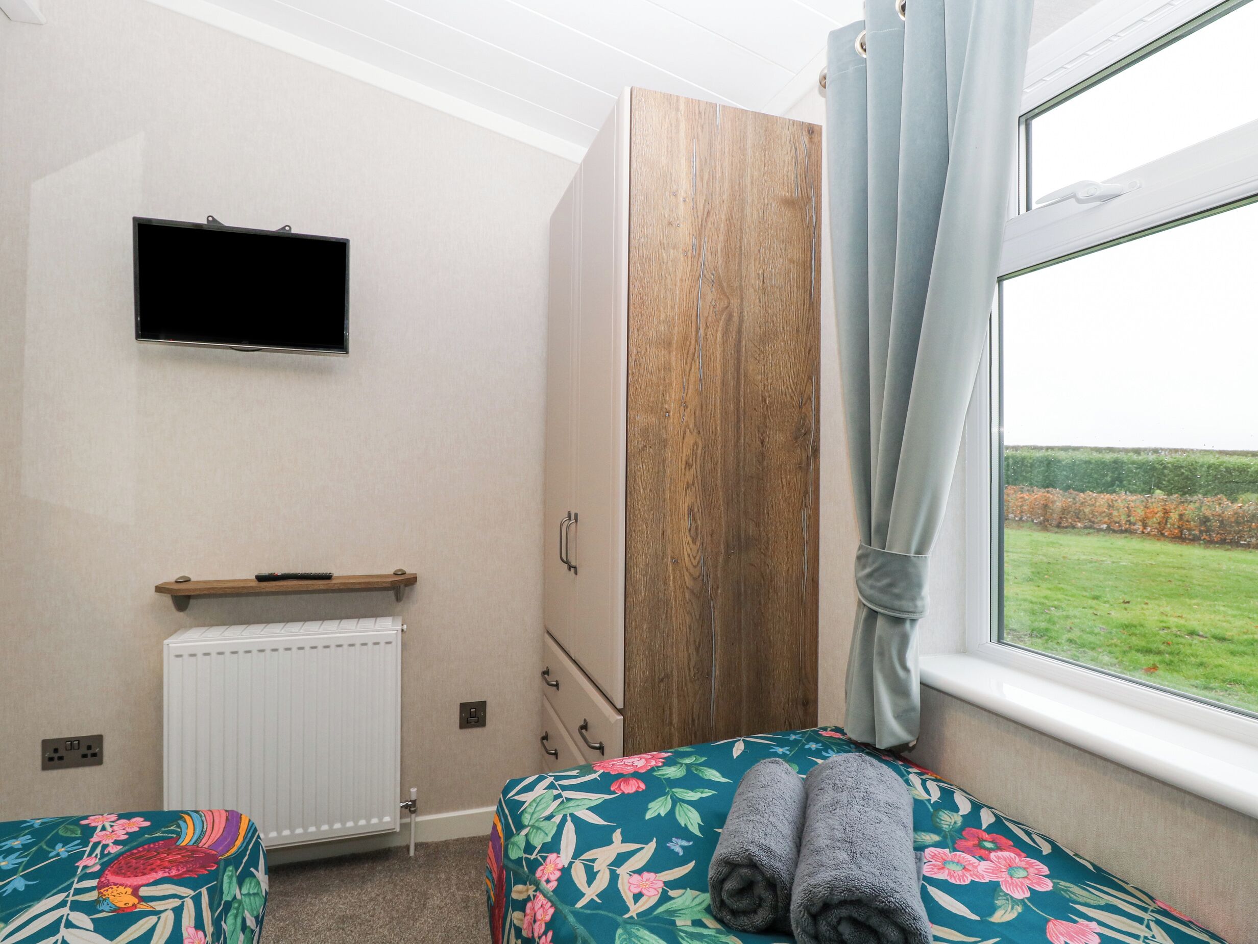 Un dormitorio con cama, armario de madera, radiador, televisor y ventana con vista a un campo.