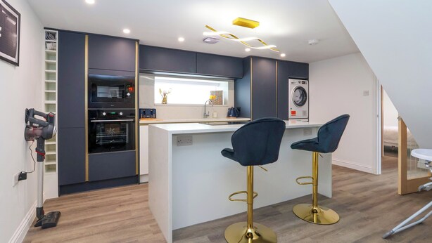A modern kitchen with a built-in oven, a washing machine, and two bar stools. - The Railway Cottage (Llandudno)