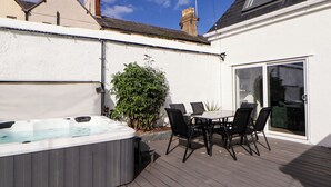 A patio with a hot tub, a table, and chairs, surrounded by a white wall and a garden. - The Railway Cottage (Llandudno)