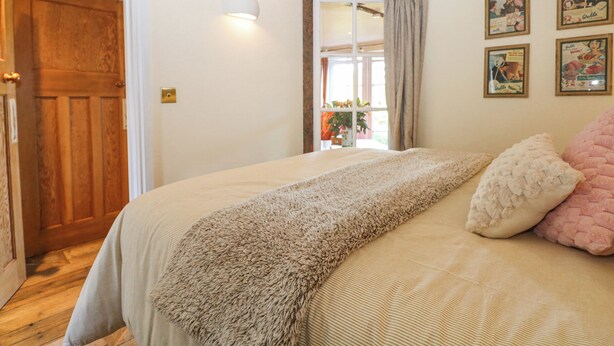 A bedroom with a wooden door, a bed with a beige and white striped bedspread, a fluffy beige throw blanket, and a pink pillow. There are framed pictures on the wall and a window with curtains. - The Bike Shed (Romsey)
