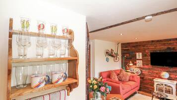 A living room with a wooden shelf holding wine glasses and a bowl, a pink sofa, a small table with a vase of flowers, and a brick wall with a mounted television.