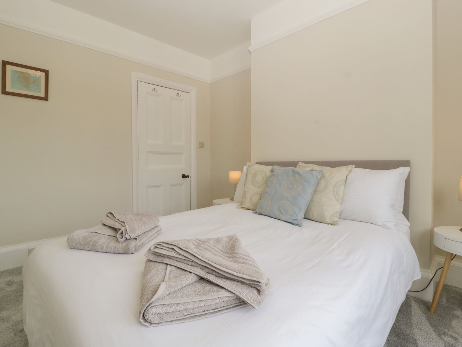 A neatly made bed with pillows and a folded towel, a white door, and a small bedside table with a lamp.