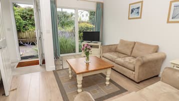 A living room with a beige sofa, a wooden coffee table, and a TV on a stand. There is a sliding glass door leading to a garden.