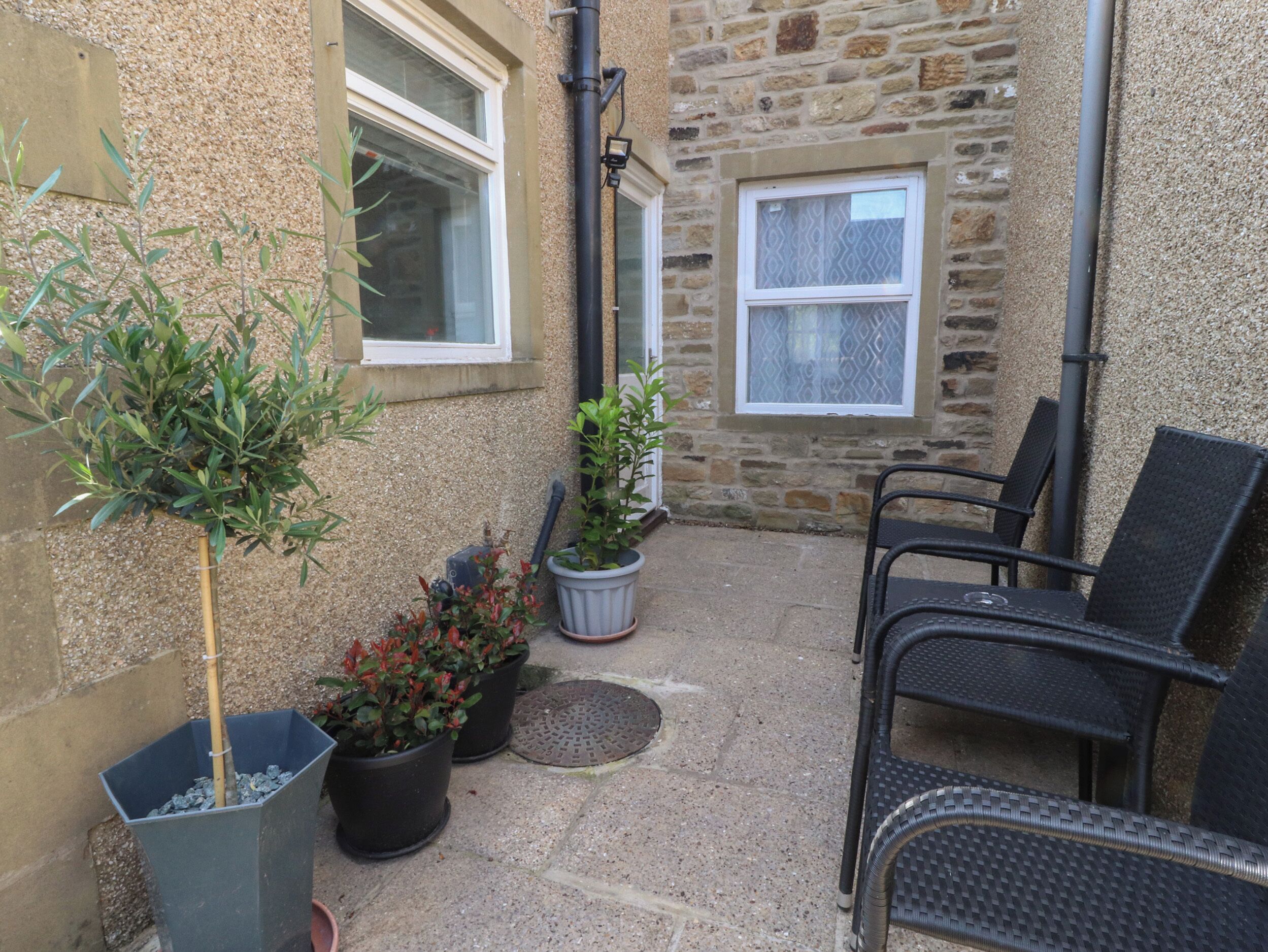 A stone building with two windows, a door, and a small outdoor seating area with chairs and potted plants.