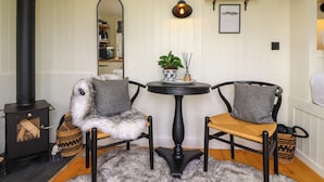 Ein gemütliches Zimmer mit einem schwarzen Kaminofen, zwei Sesseln mit grauen Kissen, einem runden Tisch mit Pflanze und Kerze sowie einem Spiegel an der Wand.