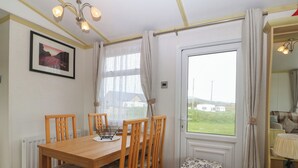 A dining area with a wooden table and chairs, a small stool, a window with a view of the countryside, and a framed picture on the wall. - Tir Glyn Caravan (Pwllheli)