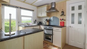 A compact kitchen with a gas stove, oven, and a small countertop. There is a window providing natural light and a view of greenery outside. - Tir Glyn Caravan (Pwllheli)