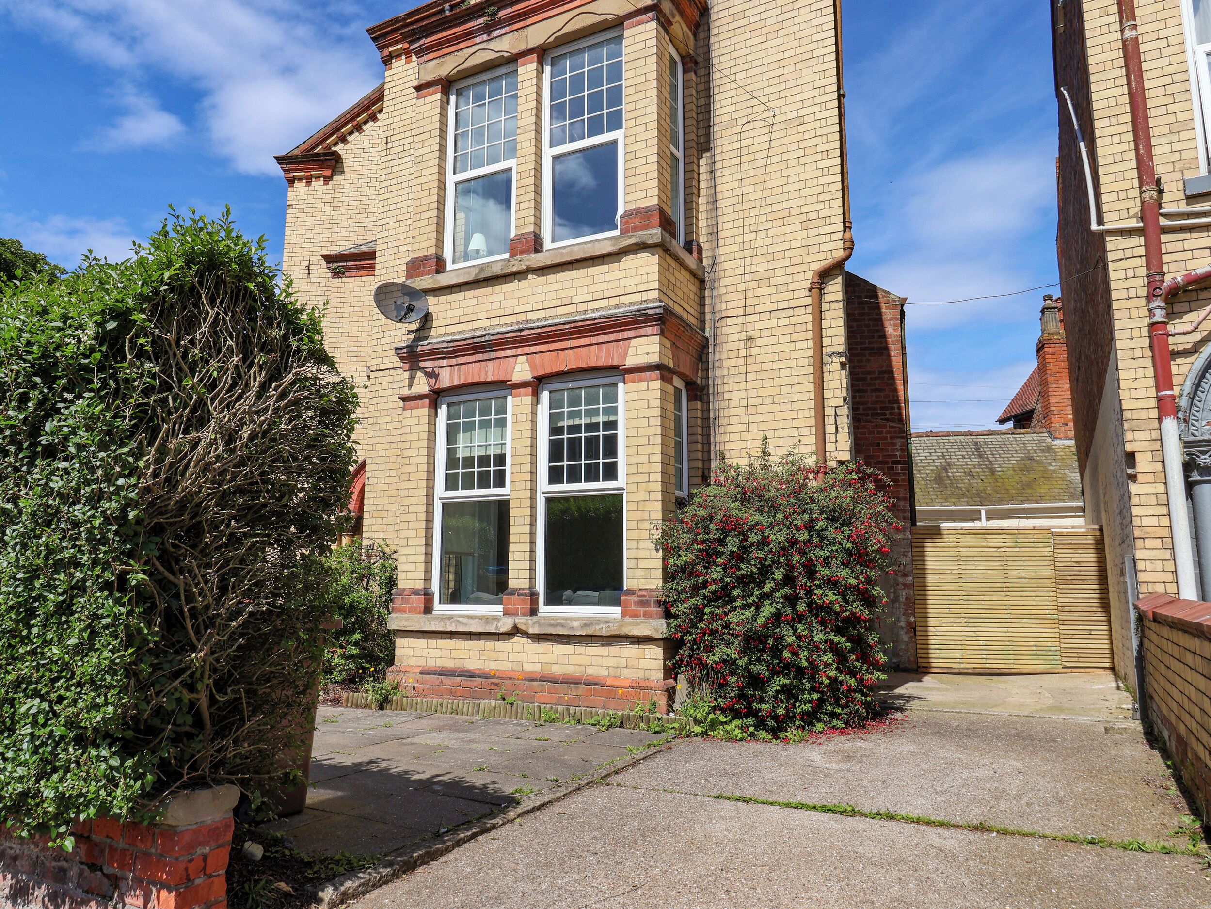 A two-story residential building with a brick facade, large windows, and a small garden area with shrubs.
