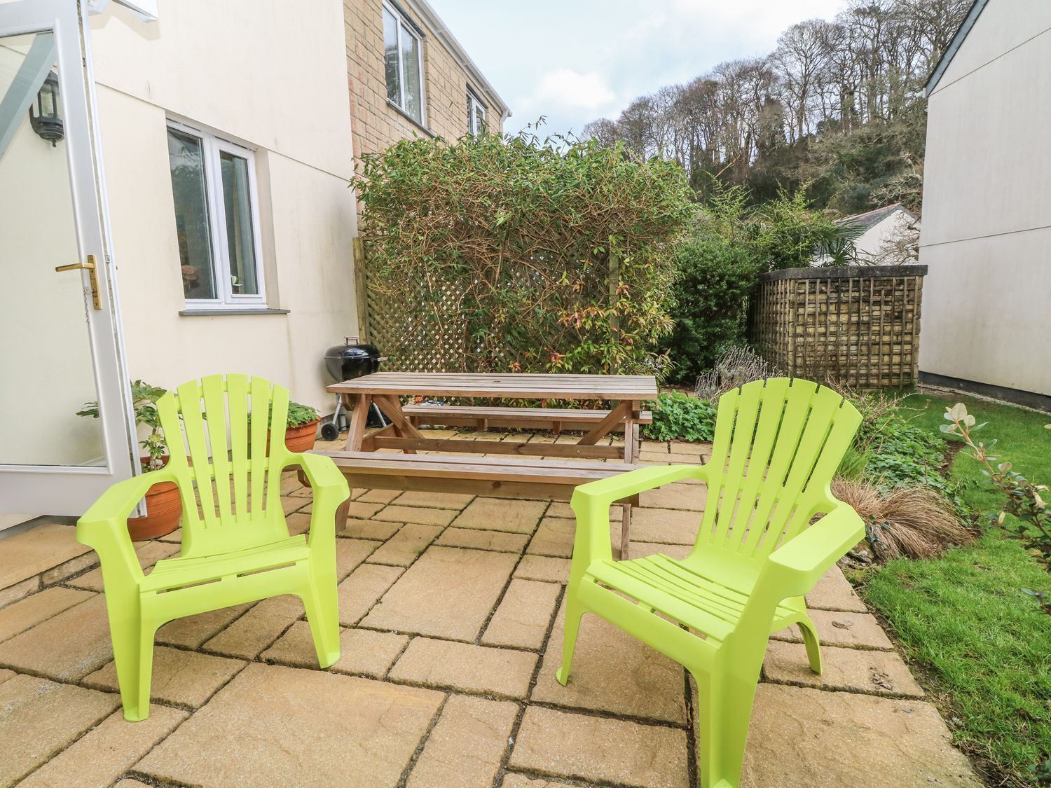 A patio with two bright green chairs and a wooden table, surrounded by a garden with a variety of plants and a fence.