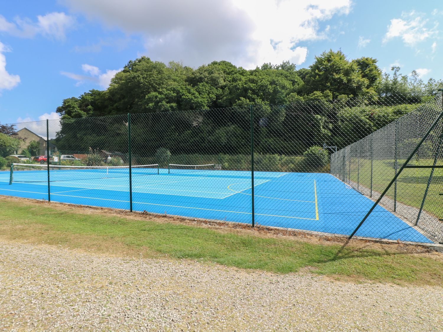 Una cancha de tenis al aire libre con una cerca de malla metálica, rodeada de césped y árboles.