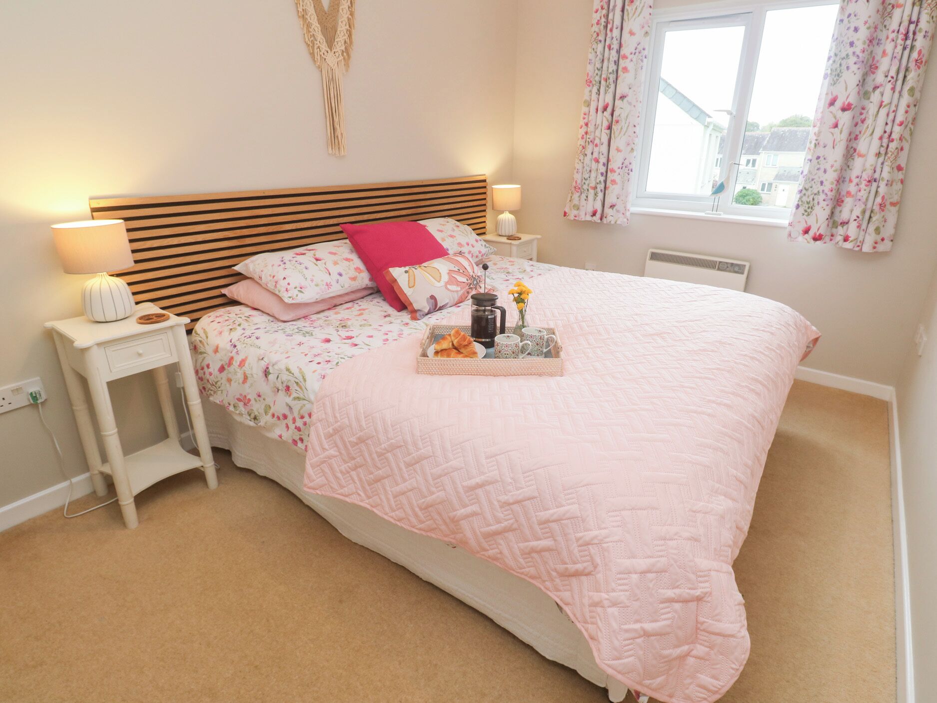 Une chambre à coucher avec une tête de lit en bois, une literie à motifs floraux, une couverture rose, des tables de chevet avec des lampes et une fenêtre ornée de rideaux à fleurs.