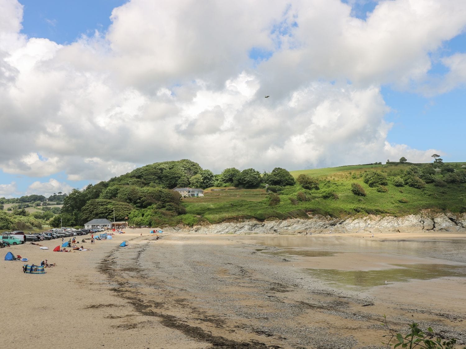 A beach with tents and cars, a grassy hill with trees, and a partly cloudy sky.