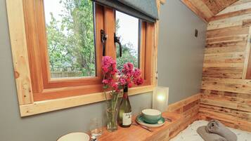 A cozy room with a wooden bed, a window with a view of greenery, a bedside table with a bowl, a wine bottle, and a vase with flowers.