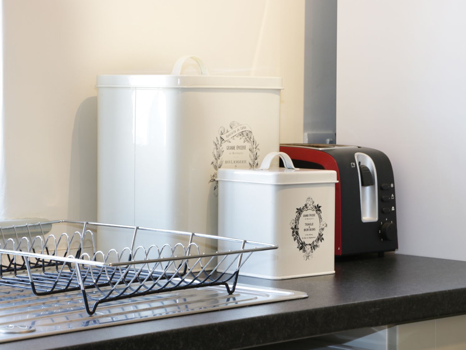 Una encimera de cocina con un escurreplatos metálico, un recipiente blanco con texto decorativo y un tostador negro.