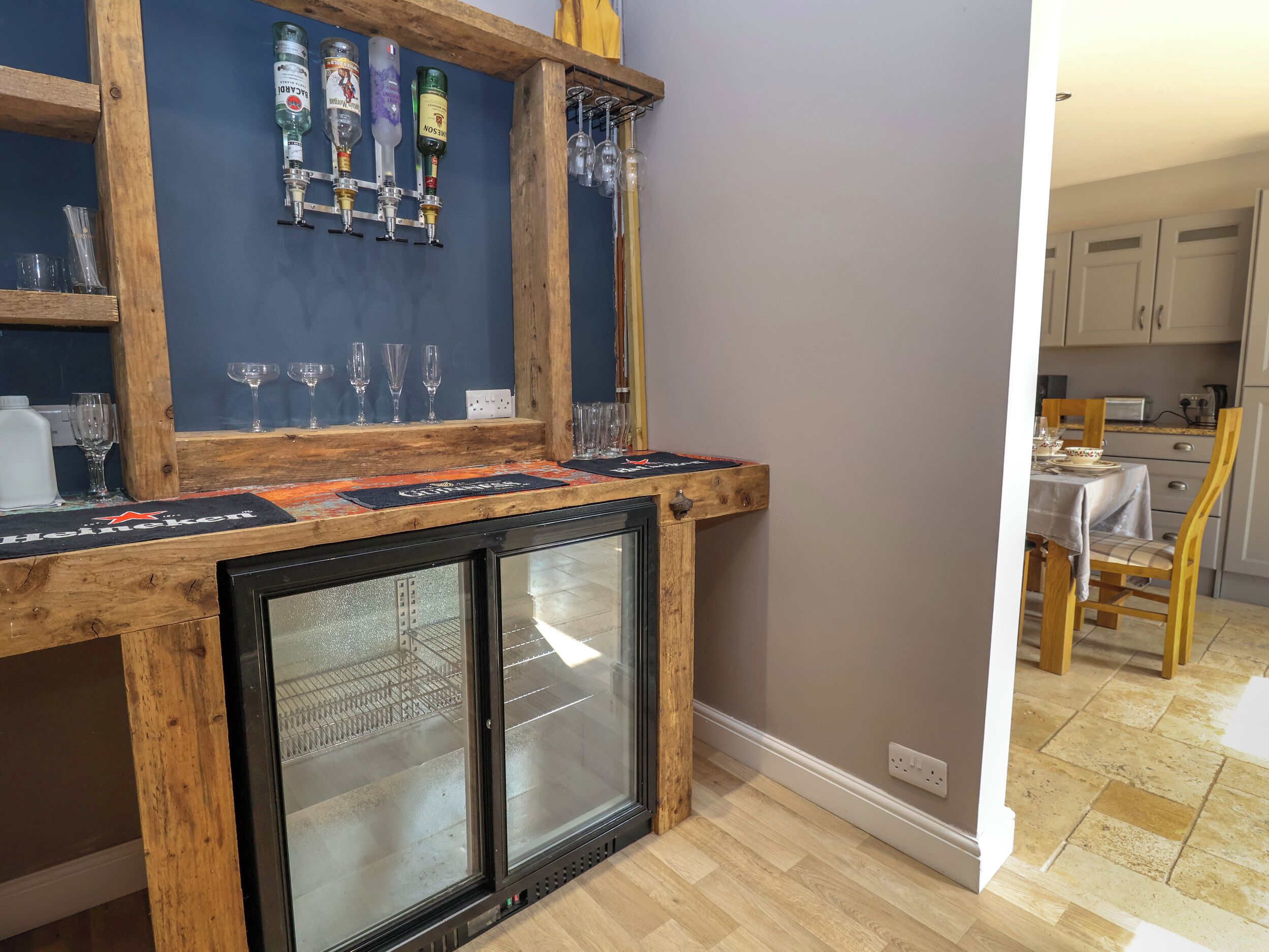 Una zona de bar con mostrador de madera, un refrigerador y una estantería para vinos con botellas y copas.