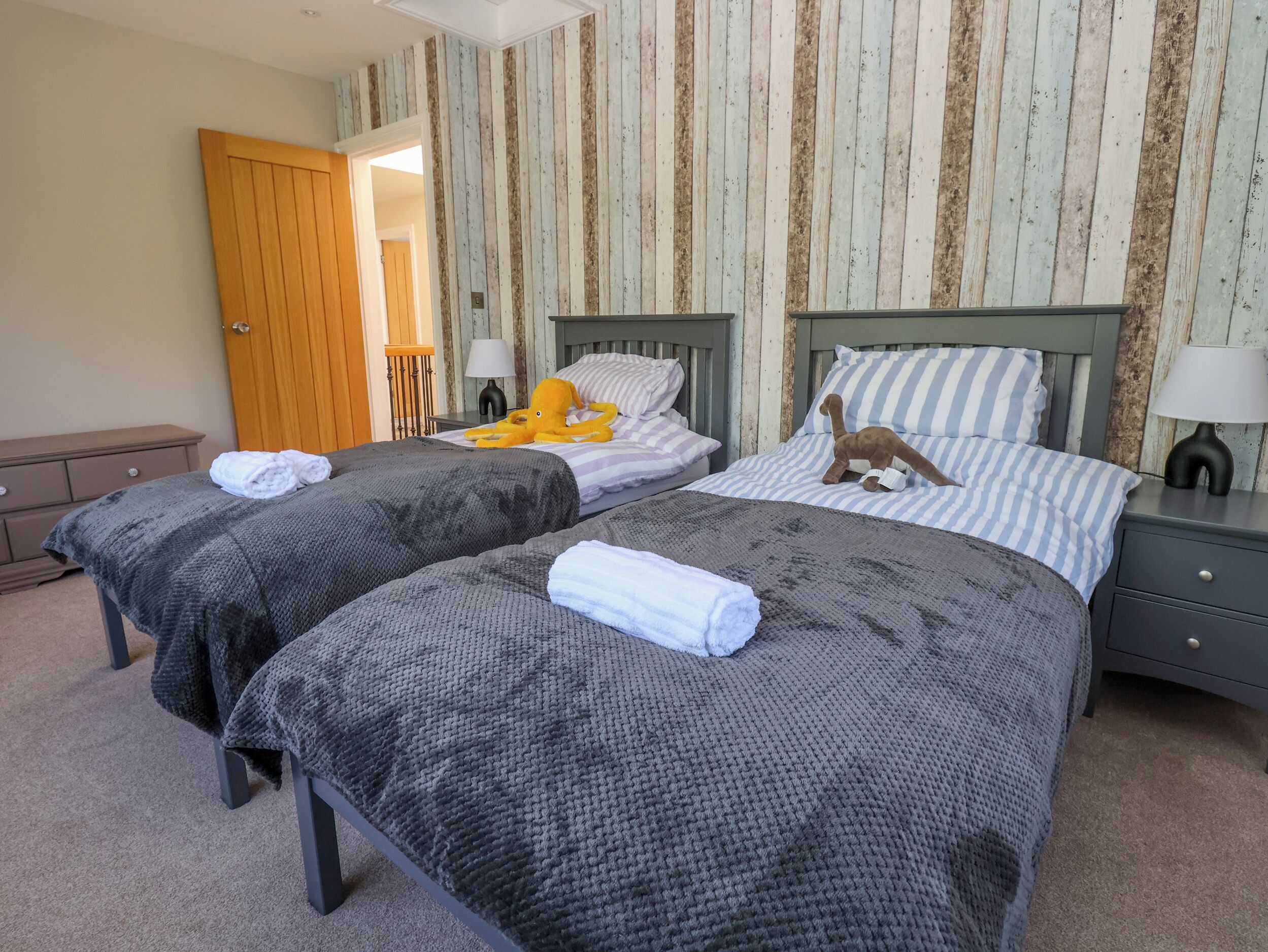 A room with two beds, each with a grey knitted blanket and a white towel. There are striped pillows and a stuffed octopus toy on one bed. The room has wooden paneling on the walls and a wooden door.
