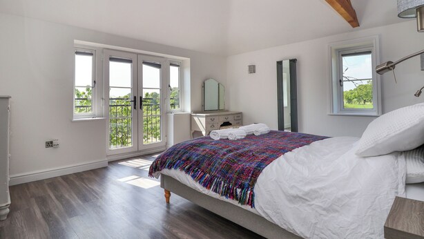 A bedroom with a large bed, a dresser, a mirror, and a wardrobe. There are two windows and a door leading to an outdoor area. - Dolly's House (Middlewich)