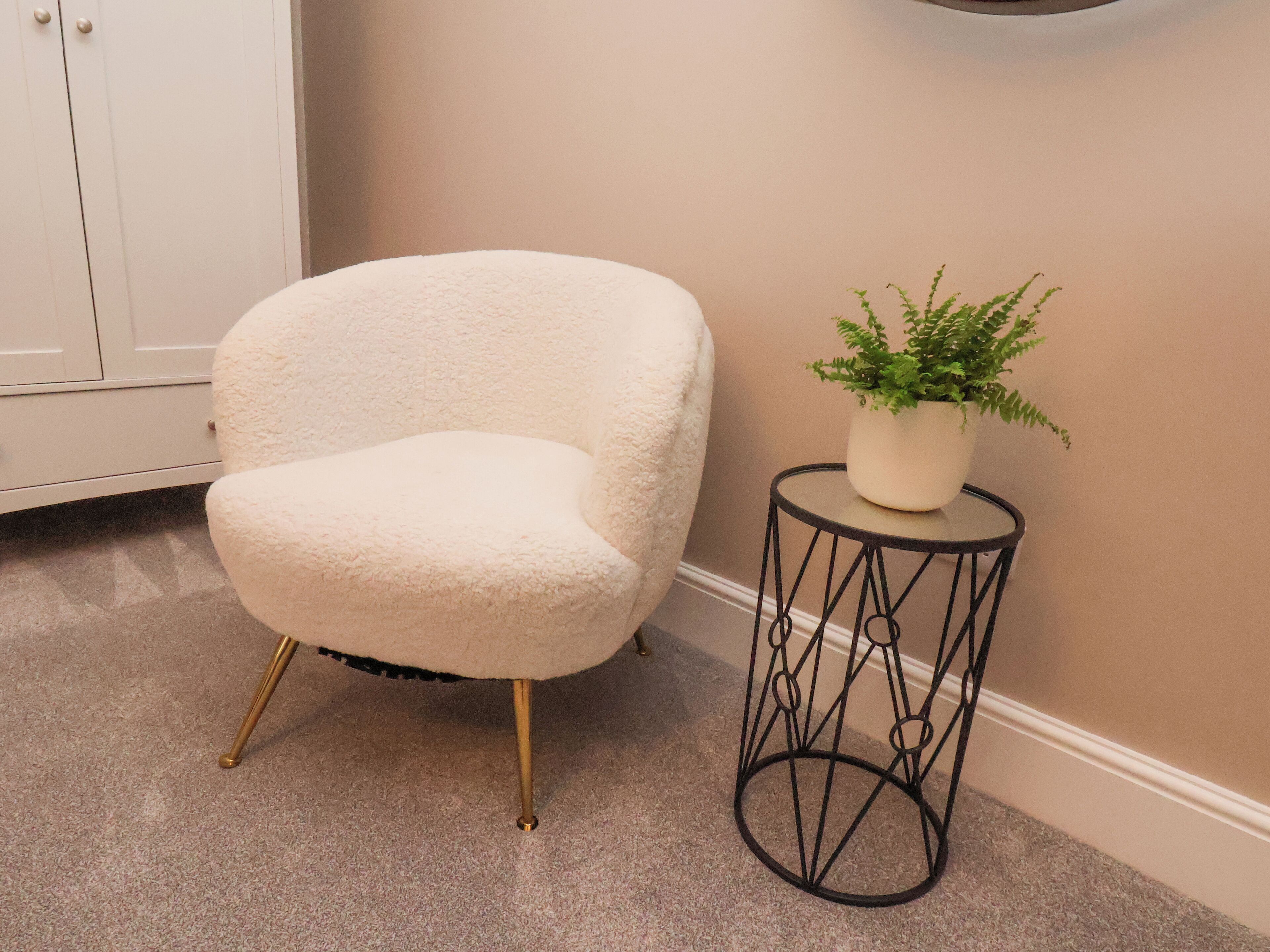 A white armchair with gold legs, a small round table with a potted plant, and a beige wall.