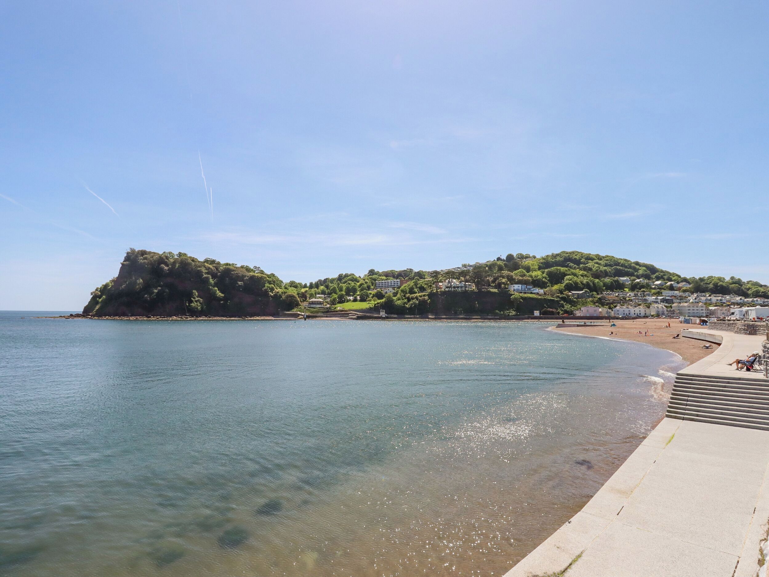 A coastal scene with a clear beach, a rocky outcrop, and buildings on a hillside.