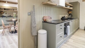 A modern kitchen with wooden beams, a dining area with a round table and chairs, and a white trash bin. - Beech Cottage - Fold Yards (Matlock)