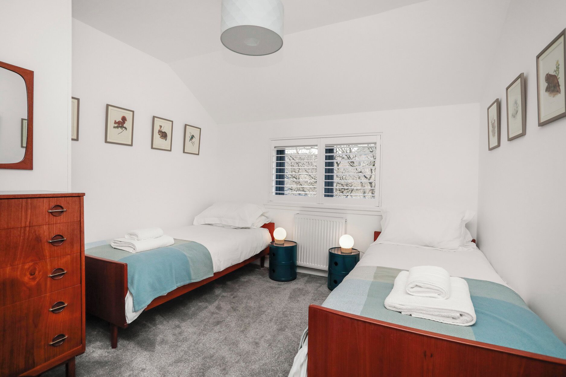 Un dormitorio con dos camas, un aparador, una ventana con persianas e imágenes enmarcadas en la pared.