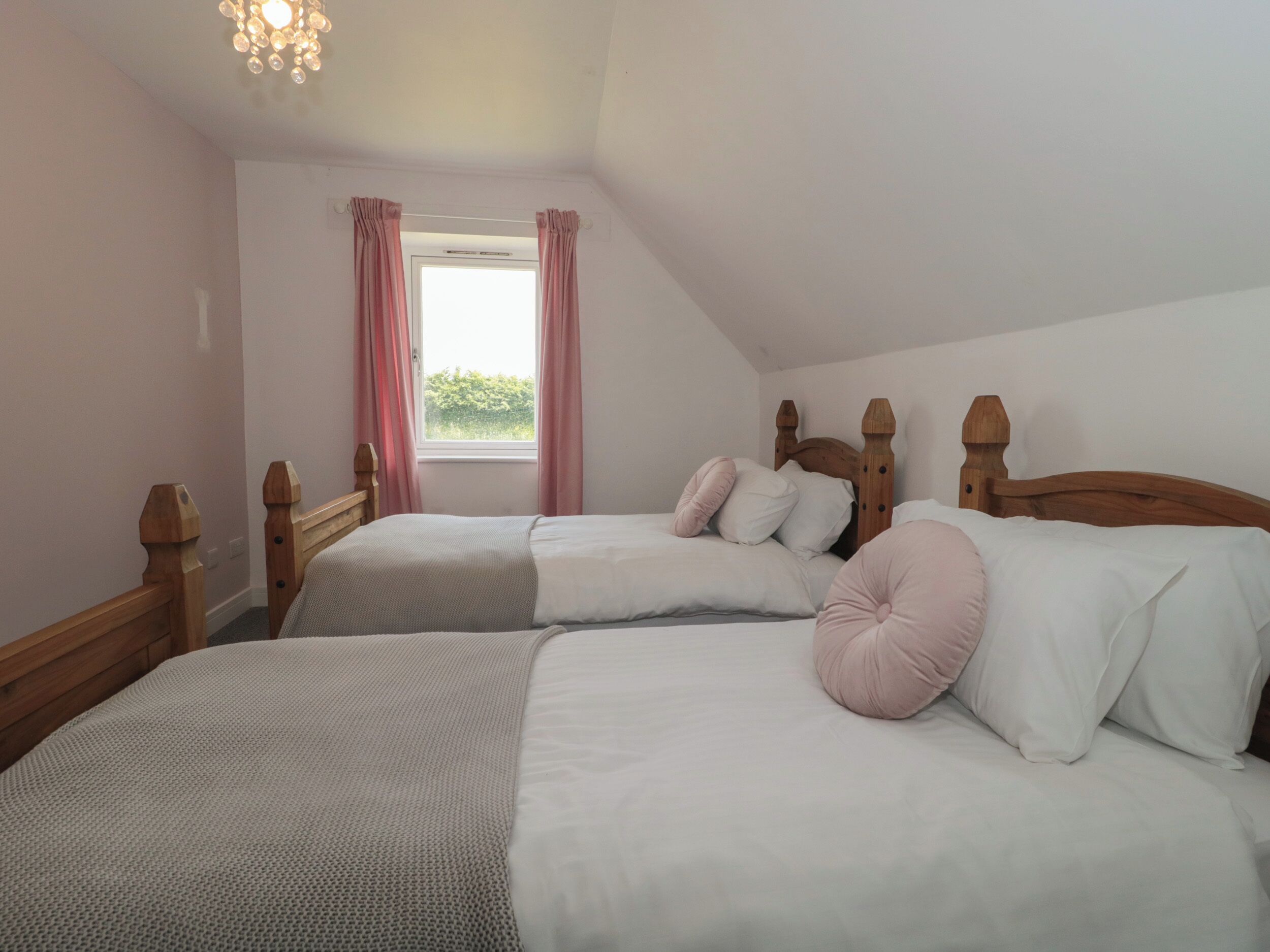 A bedroom with two single beds, wooden headboards, a window with pink curtains, and a ceiling light fixture.
