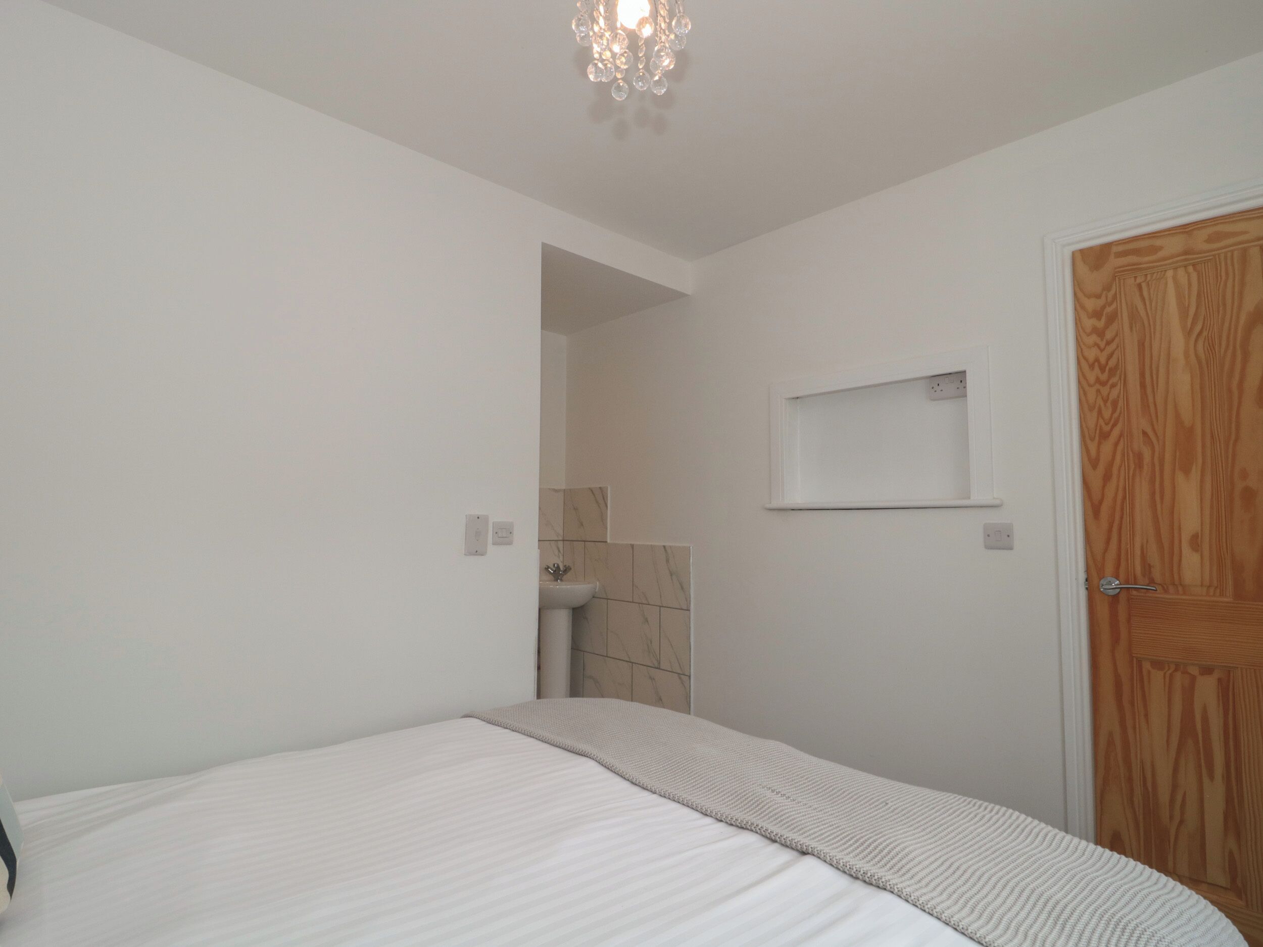 Quarto com cama, porta de madeira, prateleira pequena e banheiro visível por uma porta aberta.