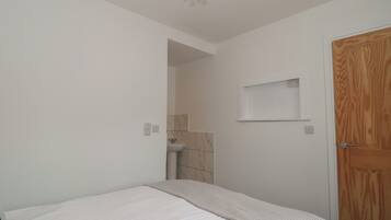 Quarto com cama, porta de madeira, prateleira pequena e banheiro visível por uma porta aberta.