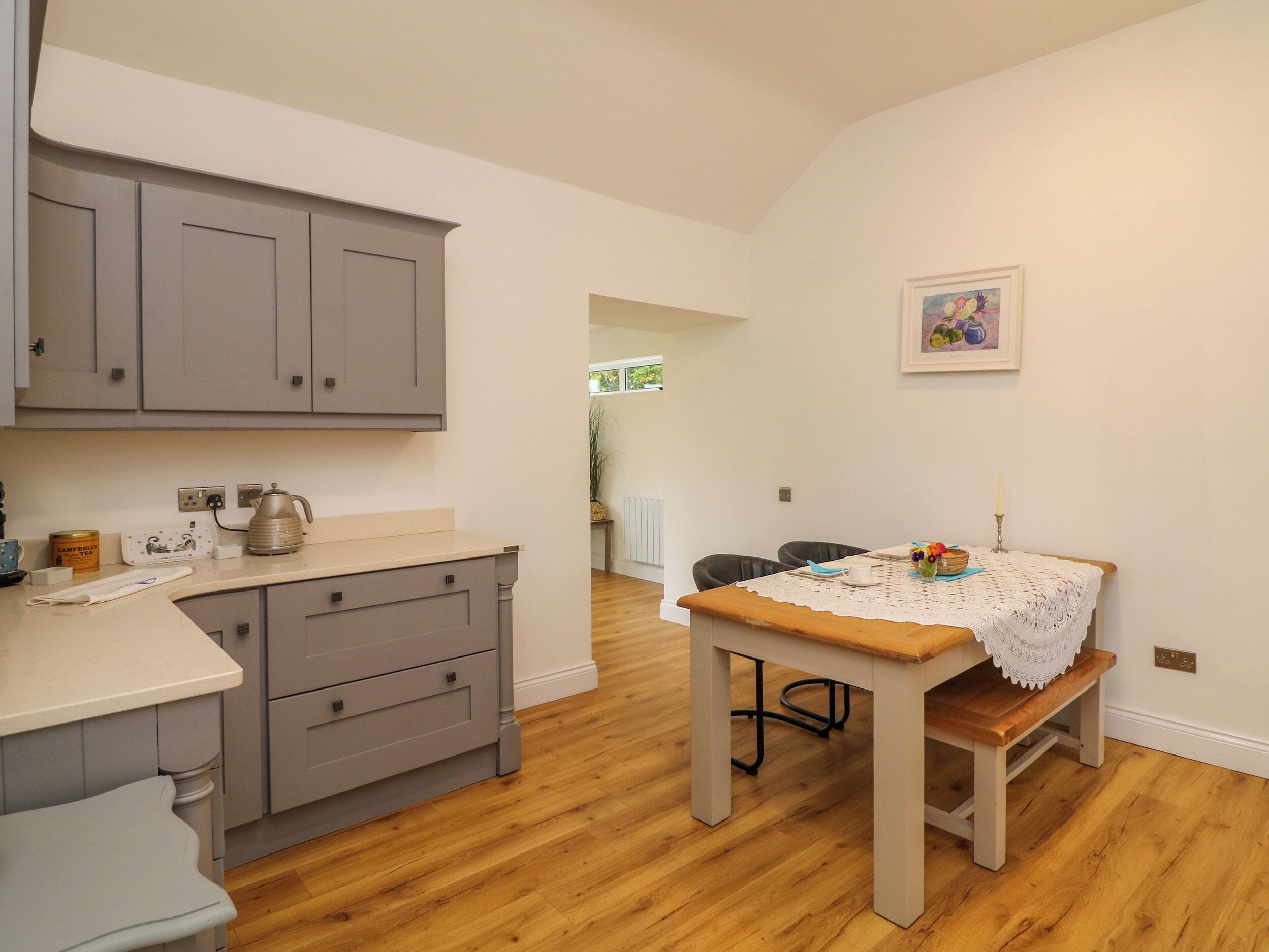 Una cocina con armarios grises, una mesa de madera con mantel de encaje y un cuadro en la pared.