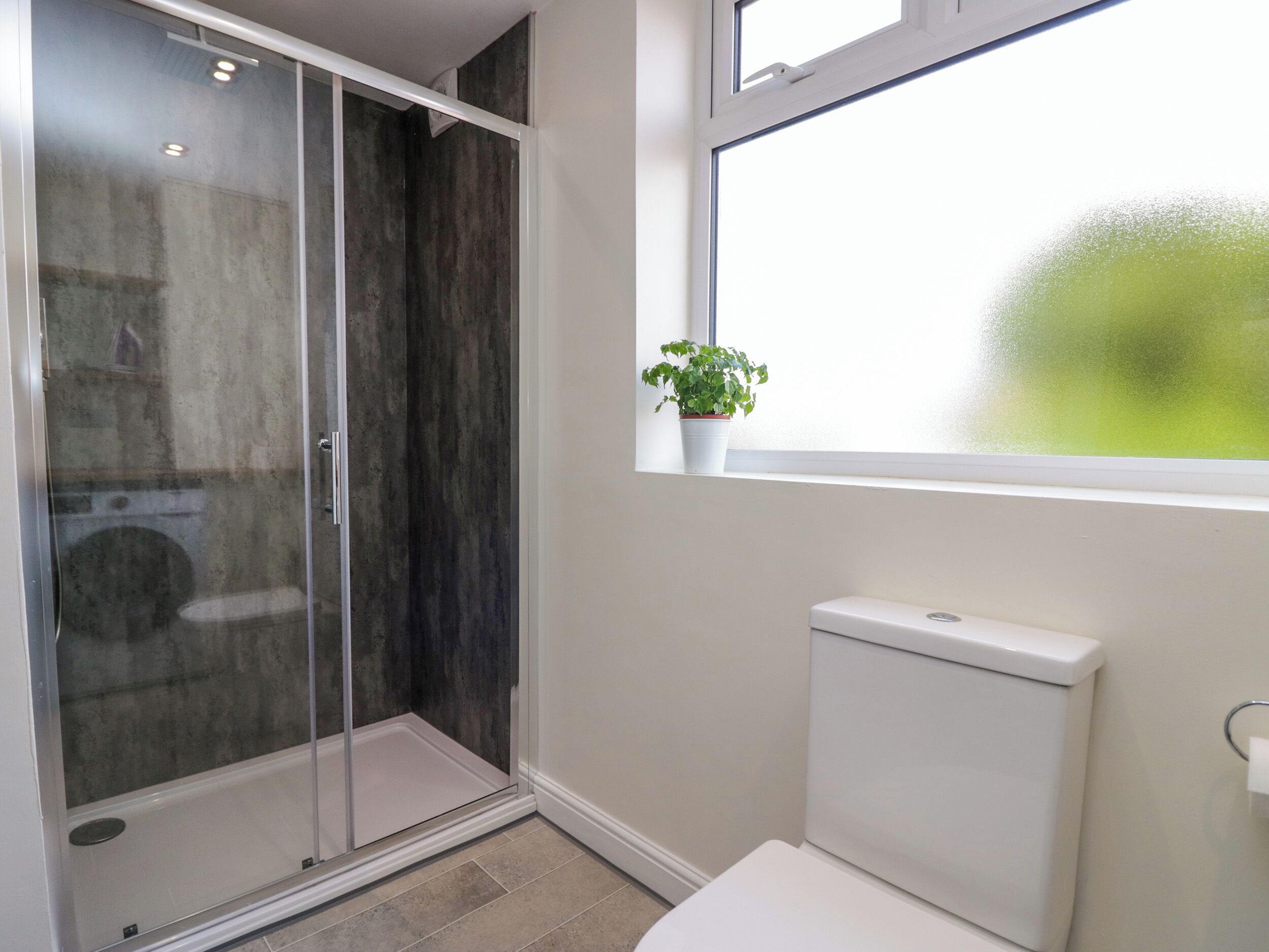 A modern bathroom with a glass shower enclosure, a toilet, and a window with a view of greenery.