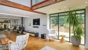 A spacious living area with a fireplace, comfortable seating, a large window with a view of the garden, and a modern kitchen in the background.