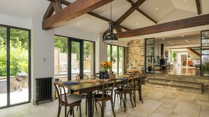A spacious dining area with a large wooden table and chairs, a stone wall, and a view of the outdoors through large windows.