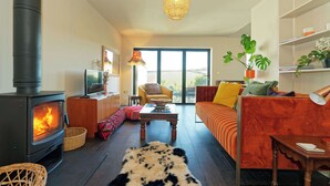 A cozy living room with a wood-burning stove, a comfortable sofa, a dining table, and a large window offering a view of the outdoors. - Kent Downs Cottage (Canterbury)