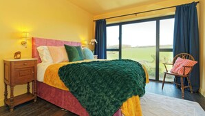 A bedroom with a large bed, a wooden nightstand, a chair, and a view of the outdoors through a large window. - Kent Downs Cottage (Canterbury)