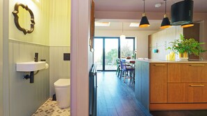 A modern kitchen with wooden cabinets, a central island, pendant lighting, and a view to an outdoor area. - Kent Downs Cottage (Canterbury)