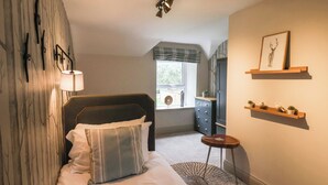 A bedroom with a bed, a bedside table, a lamp, a window, and a wall with a picture and shelves. - Low Moor (Saltburn-by-the-Sea)