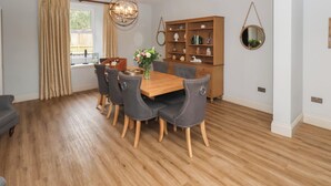 A dining room with a wooden table, six chairs, a chandelier, a bookshelf, and a window with curtains. - Low Moor (Saltburn-by-the-Sea)