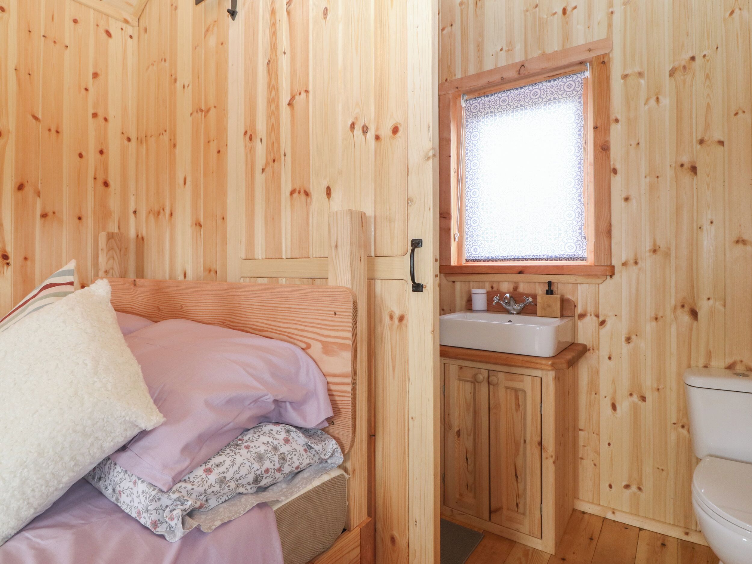 Habitación de cabaña de madera con una cama, un baño pequeño con lavamanos e inodoro, y una ventana con cortinas estampadas.