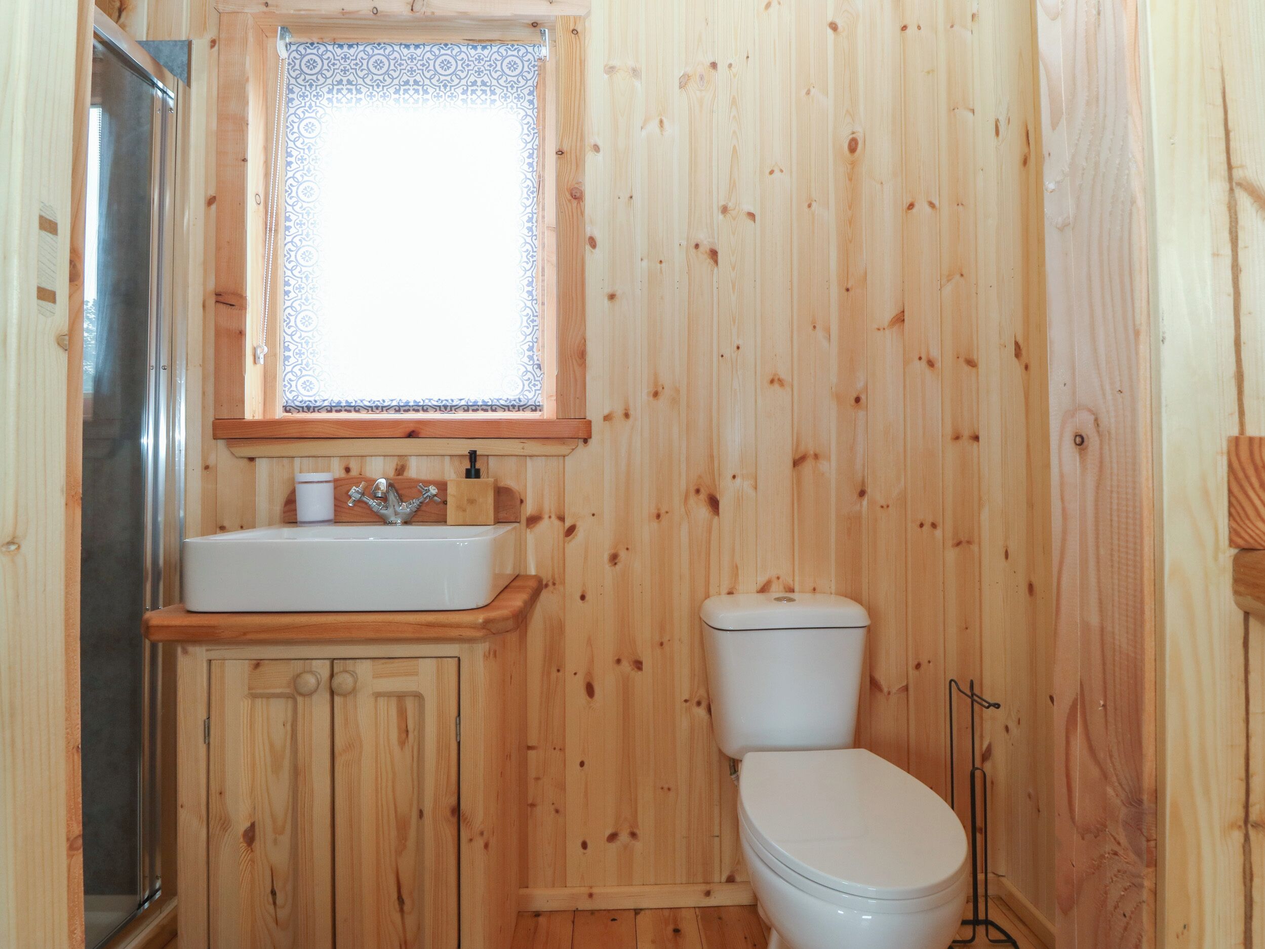 Ein Badezimmer mit Holzwandverkleidung, einem weißen Waschbecken, einer Toilette und einem Fenster mit gemusterten Vorhängen.