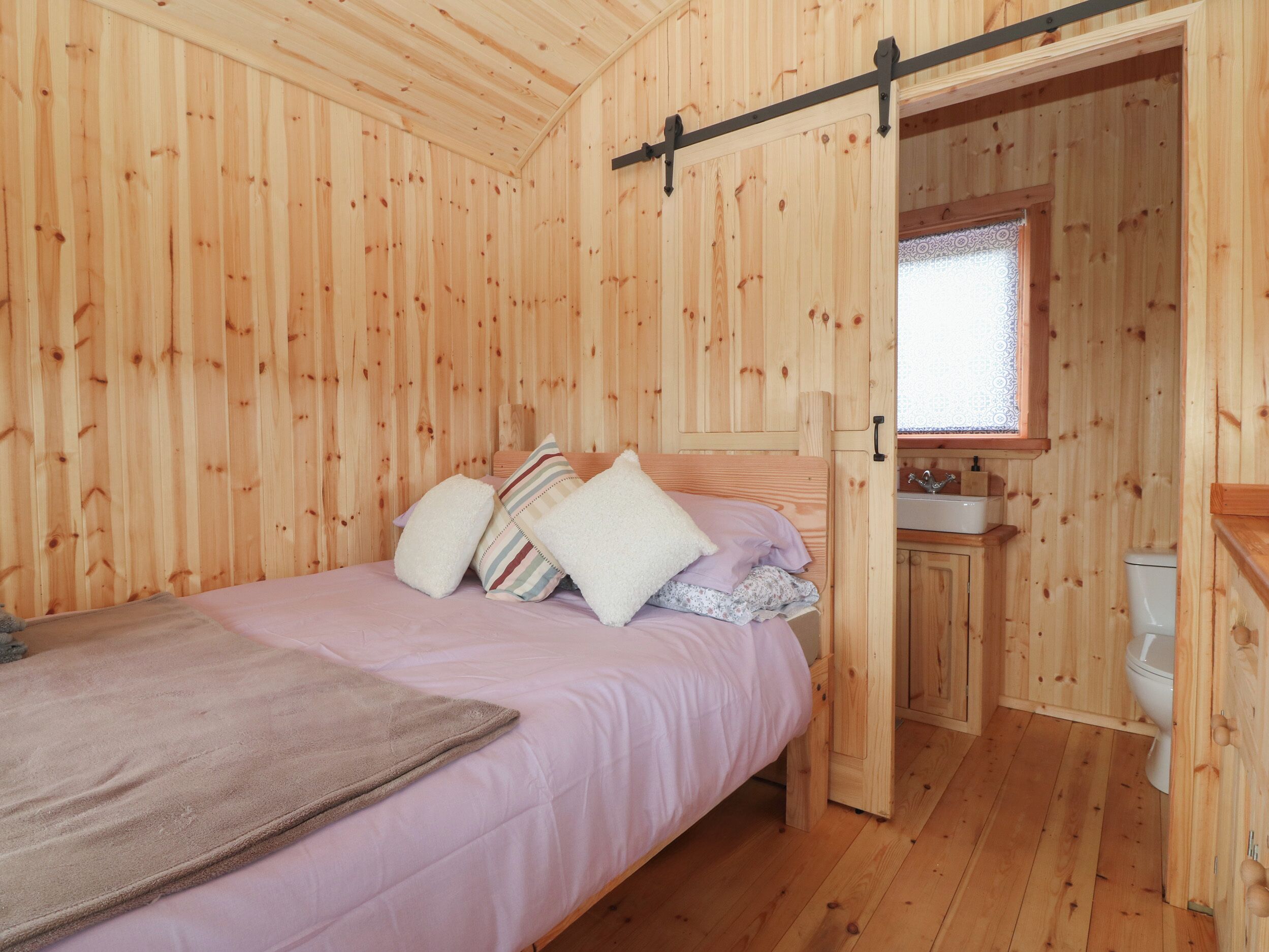 A wooden cabin room with a bed, pillows, a window, and a small bathroom.