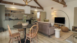 A spacious living area with a high ceiling, wooden beams, a sofa, a dining table with chairs, and a television. - Alder Cottage - Fold Yards (Matlock)