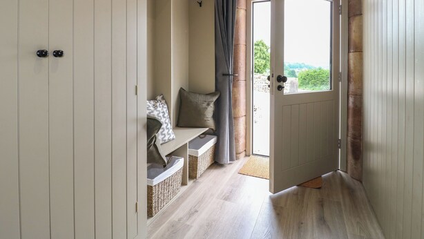 A hallway with a white door leading to a balcony, wicker baskets, and a bench with cushions. - Alder Cottage - Fold Yards (Matlock)