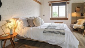A bedroom with a white brick wall, a wooden beam ceiling, a large window, a bed with white bedding, a bedside lamp, and a chair. - Alder Cottage - Fold Yards (Matlock)