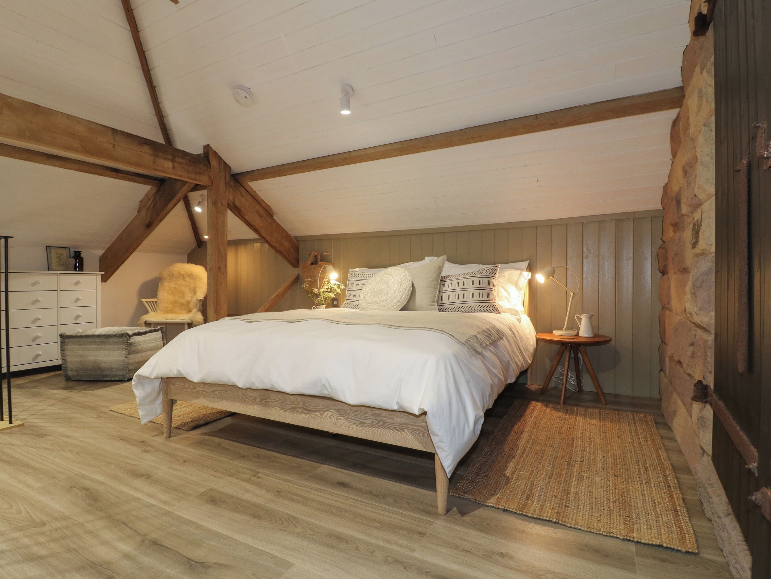 A bedroom with a wooden ceiling, a bed with white bedding, a nightstand, a chair, and a small table with a lamp.